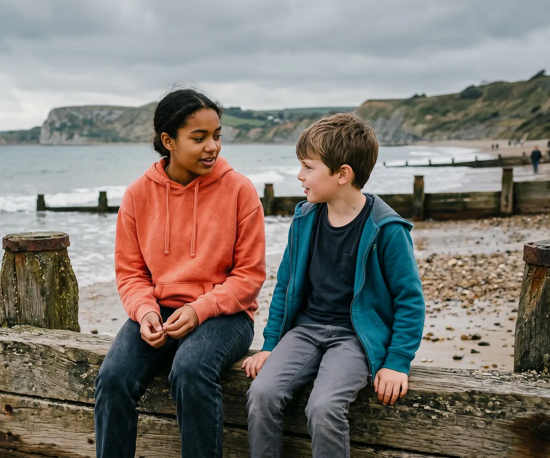 A girl in a coral hoodie and a boy in a teal jacket sit chatting on a wooden sea groyne on a beach
