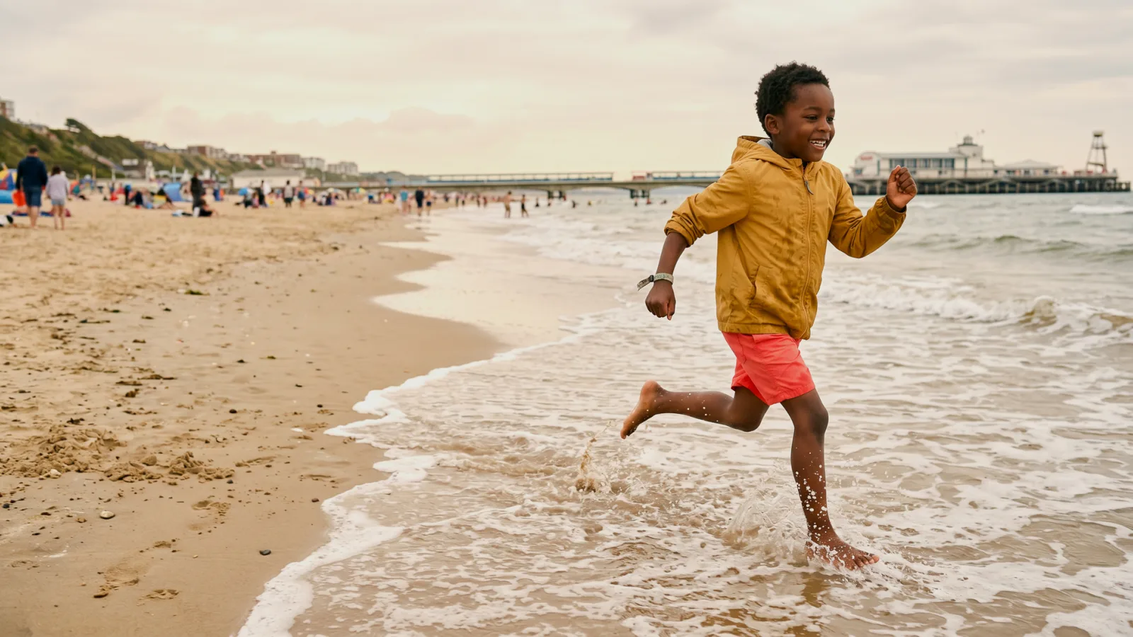 Young boy in a yellow hooded jacket and coral shorts running and laughing through the waves on a busy beach, with a pier and seafront buildings in the background