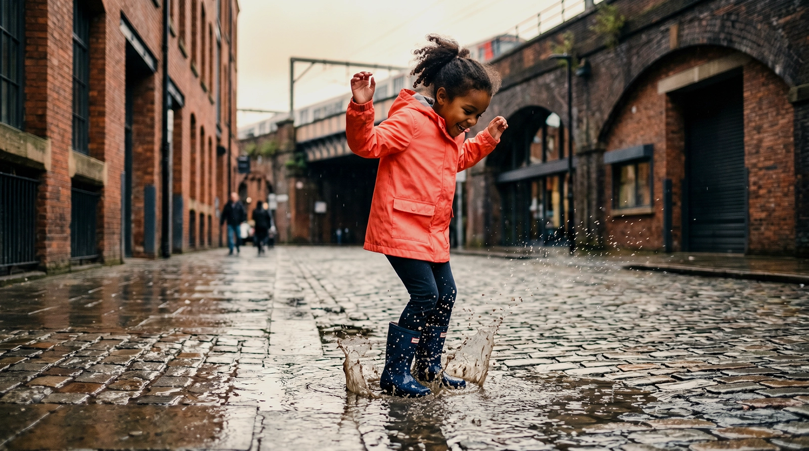 A young girl in a bright coral raincoat and blue wellies jumping joyfully in a puddle on a wet cobblestone street in Manchester, with Victorian brick arches and a railway viaduct in the background