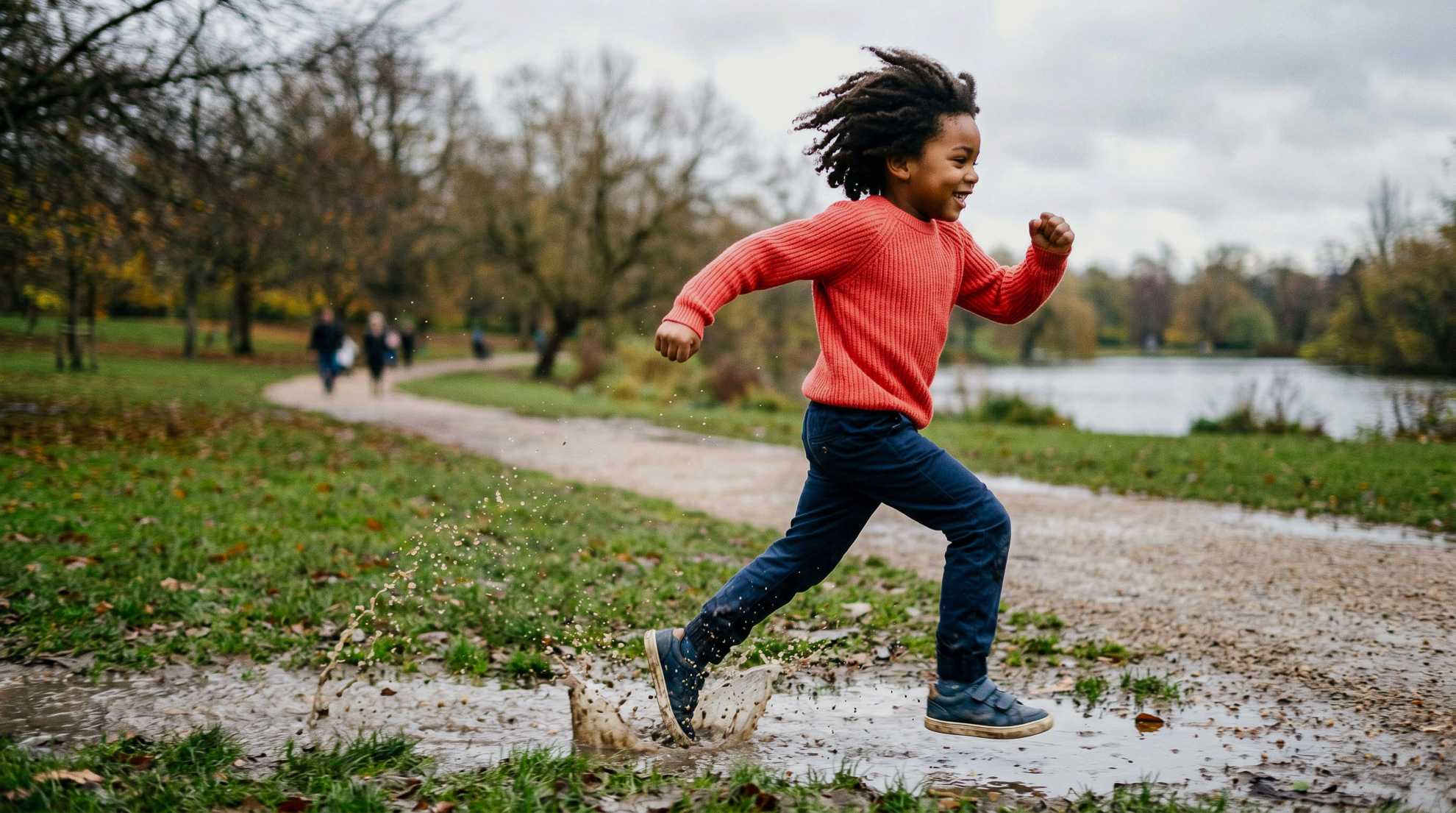 Young girl in a bright red knitted jumper running joyfully through muddy puddles on a park path beside a lake, arms pumping, with bare winter trees and a grey sky stretching behind her