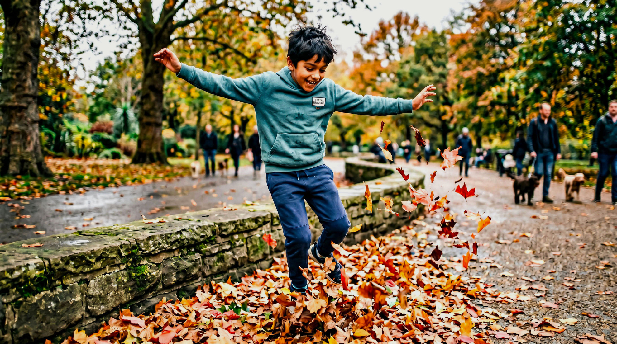 Young boy in a teal hoodie leaping joyfully through a pile of autumn leaves on a path in a Victorian park, arms outstretched and laughing, with golden trees and other families visible in the background